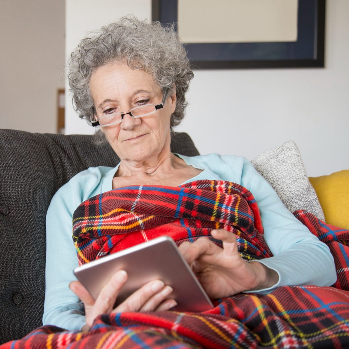 Woman on the couch wrapped in a blanket browsing her tablet